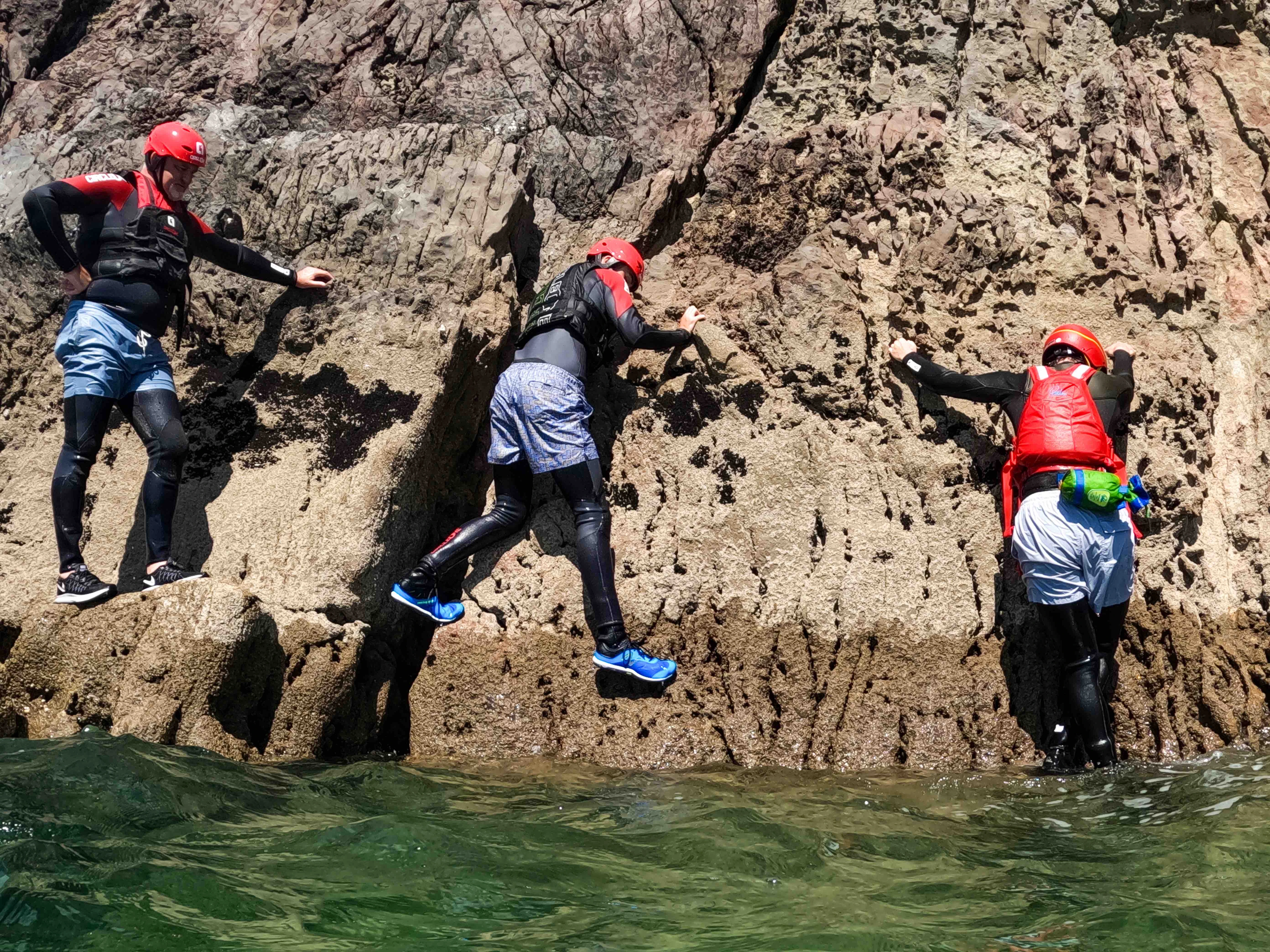A group of three people in wetsuits and helmets are coasteering in Pembrokeshire.
